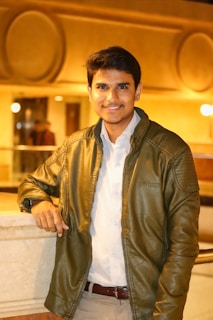 A young man is standing indoors, leaning on a marble railing. He is wearing a light-colored shirt and a green leather jacket. The background lighting is warm and there are large circular patterns on the wall behind him.