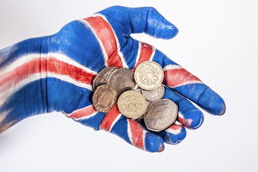 A hand painted with the blue, red, and white colors of the Union Jack is holding a collection of British coins. The vibrant paint contrasts with the metallic sheen of the coins.