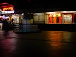 A dimly lit shopping complex with a neon sign. A sari shop displays bright red and orange fabrics in the window. People are walking past the shops, and there's a tiled courtyard in the foreground with a reflective surface.