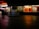 A dimly lit shopping complex with a neon sign. A sari shop displays bright red and orange fabrics in the window. People are walking past the shops, and there's a tiled courtyard in the foreground with a reflective surface.