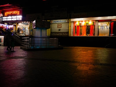 A dimly lit shopping complex with a neon sign. A sari shop displays bright red and orange fabrics in the window. People are walking past the shops, and there's a tiled courtyard in the foreground with a reflective surface.