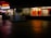 A dimly lit shopping complex with a neon sign. A sari shop displays bright red and orange fabrics in the window. People are walking past the shops, and there's a tiled courtyard in the foreground with a reflective surface.