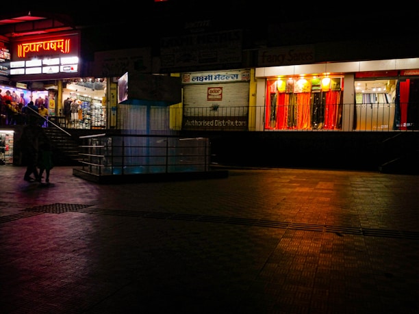 A dimly lit shopping complex with a neon sign. A sari shop displays bright red and orange fabrics in the window. People are walking past the shops, and there's a tiled courtyard in the foreground with a reflective surface.