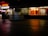 A dimly lit shopping complex with a neon sign. A sari shop displays bright red and orange fabrics in the window. People are walking past the shops, and there's a tiled courtyard in the foreground with a reflective surface.