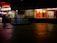 A dimly lit shopping complex with a neon sign. A sari shop displays bright red and orange fabrics in the window. People are walking past the shops, and there's a tiled courtyard in the foreground with a reflective surface.