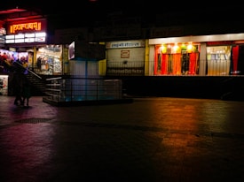 A dimly lit shopping complex with a neon sign. A sari shop displays bright red and orange fabrics in the window. People are walking past the shops, and there's a tiled courtyard in the foreground with a reflective surface.