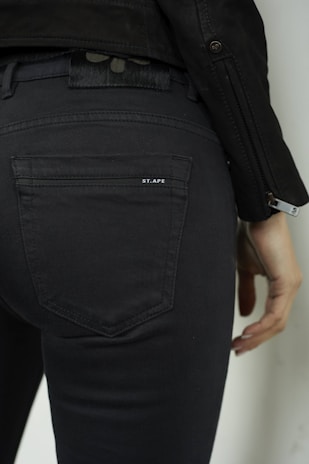 A person wearing dark-colored jeans with visible stitching, a leather jacket, and holding one hand slightly downward. The back pocket features a small logo 'ST.APE'.