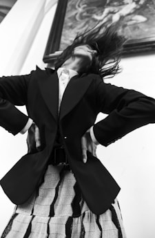 A person wearing a tailored black blazer and a patterned skirt strikes a dynamic pose with head tilted back and hair flying. The pose conveys a sense of movement and boldness. The background features an ornate framed artwork, suggesting a sophisticated setting.