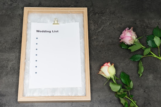 A wooden frame holds a paper titled 'Wedding List' with bullet points, placed on a dark textured surface. Two roses, one pink and one white with pink edges, lie to the right.