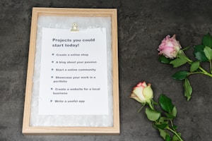 A framed piece of paper with a list of potential project ideas leaned against a dark surface. Next to it, two roses, one pink and one cream with pink edges, lay on the surface.