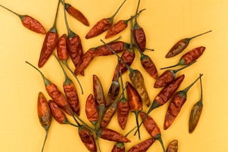 A collection of dried red chili peppers is scattered across a bright yellow background. The peppers vary in size and have long green stems attached, creating a contrasting color scheme. The texture of the chili skins is wrinkled and rough.