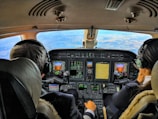 Two pilots are seated in the cockpit of an aircraft, surrounded by multiple displays and control panels. The view out the windshield shows a clear sky and expansive landscape below. Both pilots are wearing headsets, indicating active communication.