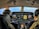 Two pilots are seated in the cockpit of an aircraft, surrounded by multiple displays and control panels. The view out the windshield shows a clear sky and expansive landscape below. Both pilots are wearing headsets, indicating active communication.