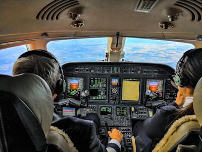 Two pilots are seated in the cockpit of an aircraft, surrounded by multiple displays and control panels. The view out the windshield shows a clear sky and expansive landscape below. Both pilots are wearing headsets, indicating active communication.