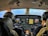 Two pilots are seated in the cockpit of an aircraft, surrounded by multiple displays and control panels. The view out the windshield shows a clear sky and expansive landscape below. Both pilots are wearing headsets, indicating active communication.