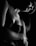 A black and white photograph capturing an artistic composition of a partially lit figure. The lighting emphasizes the contours of the shoulder and arm, creating a dramatic contrast. The texture of the skin and a hint of a strapless top are visible. The overall composition is intimate and moody.