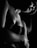 A black and white photograph capturing an artistic composition of a partially lit figure. The lighting emphasizes the contours of the shoulder and arm, creating a dramatic contrast. The texture of the skin and a hint of a strapless top are visible. The overall composition is intimate and moody.