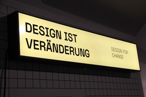 A rectangular, illuminated sign displays the phrase 'DESIGN IST VER&Auml;NDERUNG' on the left side and 'DESIGN FOR CHANGE' on the right side. The sign has a yellow background with black text and is mounted against a tiled wall.