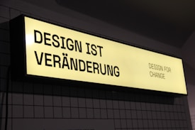 A rectangular, illuminated sign displays the phrase 'DESIGN IST VERÄNDERUNG' on the left side and 'DESIGN FOR CHANGE' on the right side. The sign has a yellow background with black text and is mounted against a tiled wall.