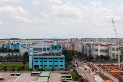 A cityscape featuring numerous high-rise residential buildings with colorful facades. In the foreground, there is an open area with parked buses surrounded by greenery. To the right, a construction site with a tall crane is visible, indicating ongoing development. The sky is partly cloudy, contributing to a calm urban atmosphere.