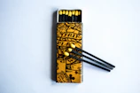 An open matchbox with a yellow and black artistic design on the cover. Inside, matches with black stems and yellow heads are visible. Some matches are positioned outside the box, resting on the surface.