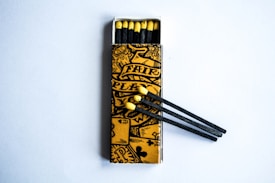 An open matchbox with a yellow and black artistic design on the cover. Inside, matches with black stems and yellow heads are visible. Some matches are positioned outside the box, resting on the surface.