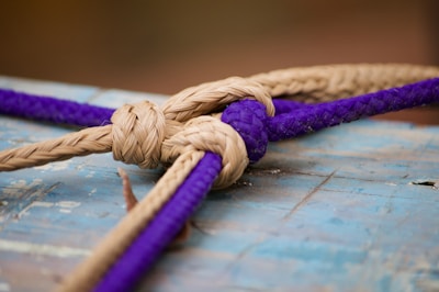 A close-up shot of a knot securing two ropes, one tan and one purple, against a weathered wooden surface with a blueish tint. The ropes are intricately weaved together in a detailed knot.