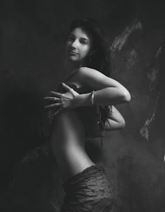 A black and white artistic photograph of a woman standing in profile with her head turned towards the camera. Her long hair cascades down her back, and she wears a stylish outfit. The lighting accentuates the contours of her body, creating a dramatic effect against a textured, dark backdrop.