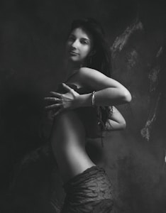 A black and white artistic photograph of a woman standing in profile with her head turned towards the camera. Her long hair cascades down her back, and she wears a stylish outfit. The lighting accentuates the contours of her body, creating a dramatic effect against a textured, dark backdrop.