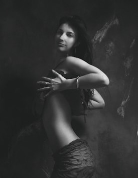 A black and white artistic photograph of a woman standing in profile with her head turned towards the camera. Her long hair cascades down her back, and she wears a stylish outfit. The lighting accentuates the contours of her body, creating a dramatic effect against a textured, dark backdrop.