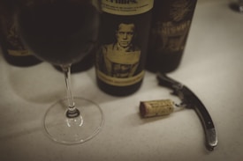A wine glass filled with red wine is placed on a light-colored surface alongside a dark wine bottle featuring a vintage-style label with a black and white portrait. Nearby, a corkscrew and a wine cork lie on the table.