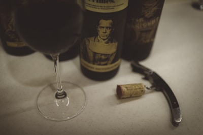 A wine glass filled with red wine is placed on a light-colored surface alongside a dark wine bottle featuring a vintage-style label with a black and white portrait. Nearby, a corkscrew and a wine cork lie on the table.