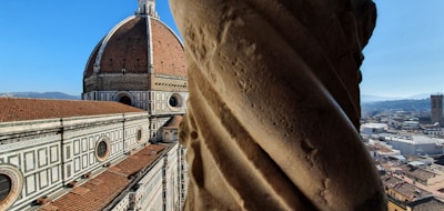 A view featuring the Florence Cathedral with its iconic dome in the background and a large, detailed stone column in the foreground. The cityscape spreads out to the right, under a clear blue sky, with distant hills visible.