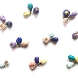 Clusters of small, colorful ceramic flower pendants arranged on a light background. Each cluster contains delicate blooms in various shades such as blue, purple, pink, and yellow, with some petals having a metallic accent.