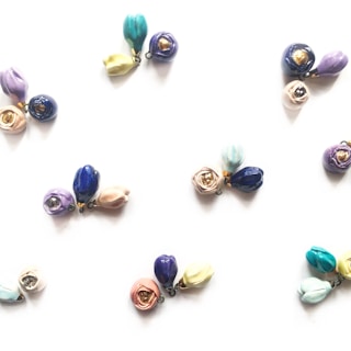 Clusters of small, colorful ceramic flower pendants arranged on a light background. Each cluster contains delicate blooms in various shades such as blue, purple, pink, and yellow, with some petals having a metallic accent.