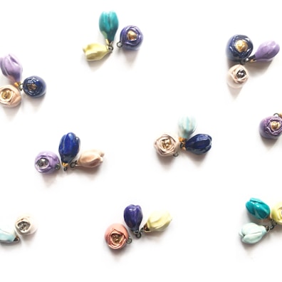 Clusters of small, colorful ceramic flower pendants arranged on a light background. Each cluster contains delicate blooms in various shades such as blue, purple, pink, and yellow, with some petals having a metallic accent.