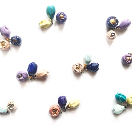 Clusters of small, colorful ceramic flower pendants arranged on a light background. Each cluster contains delicate blooms in various shades such as blue, purple, pink, and yellow, with some petals having a metallic accent.