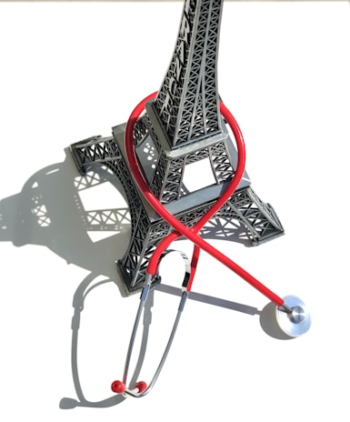 A small model of the Eiffel Tower is intertwined with a red stethoscope, casting a shadow on a white background.