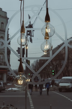 Glass light bulbs hang against a backdrop of a busy urban street. The bulbs are turned on and feature a decorative design in the filament. The image also includes a geometric pattern on the window through which the street and pedestrians are visible.