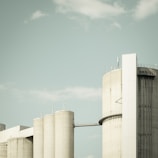 A set of large cylindrical industrial buildings, possibly silos or storage tanks, stand beneath a pale, cloudy sky. The structures are made of concrete and have a very utilitarian design with minimal decoration.