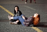 Two women are sitting and lying on a cobblestone street. One woman with dark hair and a black jacket is seated and holding a mobile phone, looking at the other woman with a smile. The other woman with long red hair is lying on her back, with her legs resting on the seated woman's lap, and appears to be laughing. They are both dressed casually in jeans and jackets. There is a yellow line on the street next to them, and it appears to be nighttime or early evening due to the dim lighting.