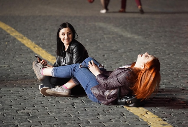 Two women are sitting and lying on a cobblestone street. One woman with dark hair and a black jacket is seated and holding a mobile phone, looking at the other woman with a smile. The other woman with long red hair is lying on her back, with her legs resting on the seated woman's lap, and appears to be laughing. They are both dressed casually in jeans and jackets. There is a yellow line on the street next to them, and it appears to be nighttime or early evening due to the dim lighting.