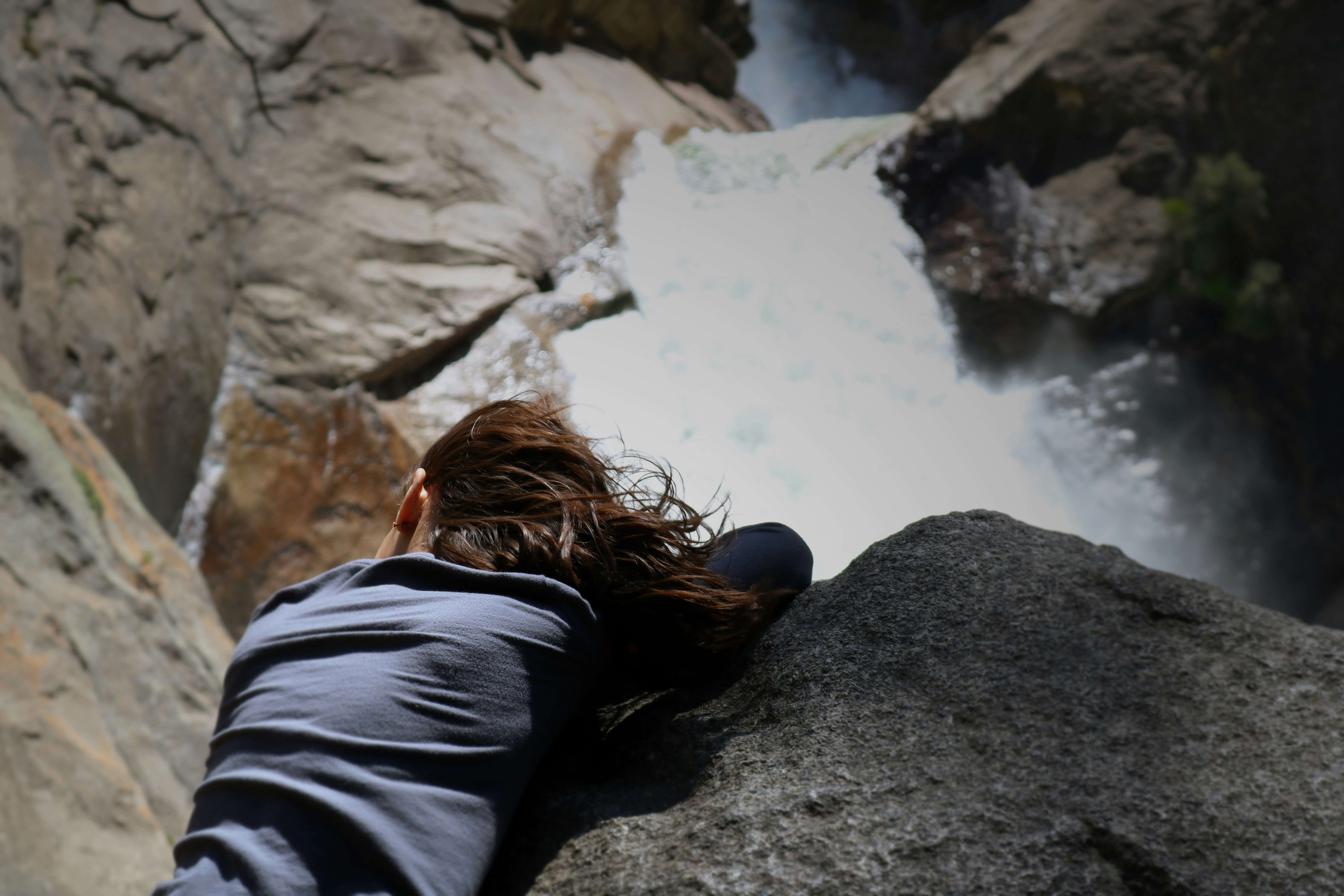 woman in black jacket sitting on rock near river during daytime, Feeling the wind in her hair from the waterfall fall.