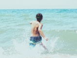 A child runs into the ocean waves, creating splashes of water as they move. The scene captures a sense of freedom and playfulness, set against the backdrop of a vast, blue sea.