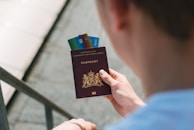 A person is holding a Dutch passport with several credit cards inserted between its pages. The passport has an emblem with a lion, and the text is written in gold. The person is wearing a light-colored shirt, and the background is slightly blurred, showing a staircase.