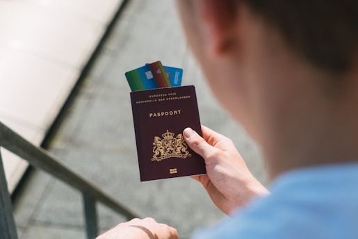 A person is holding a Dutch passport with several credit cards inserted between its pages. The passport has an emblem with a lion, and the text is written in gold. The person is wearing a light-colored shirt, and the background is slightly blurred, showing a staircase.