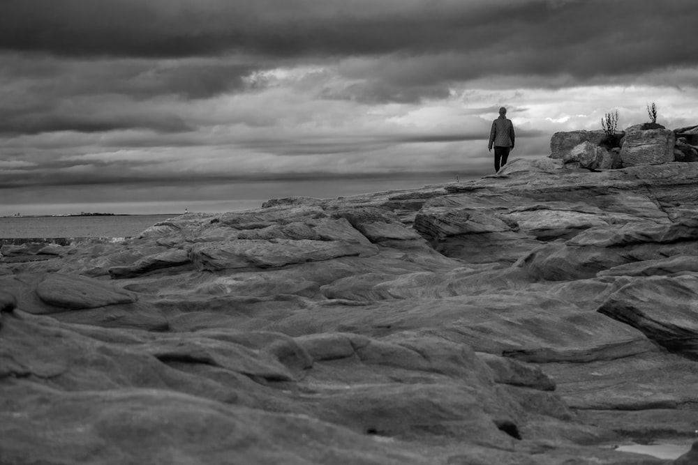 grayscale photo of man standing on rock formation near sea