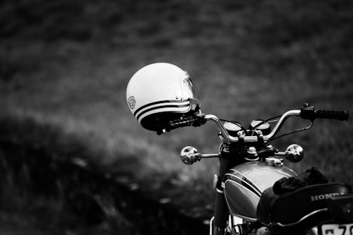 A black and white photograph of a classic motorcycle with a vintage design, positioned in the foreground. A helmet with a retro style and stripe pattern is placed on the right handlebar. The background is blurred, showcasing a grassy field, which adds depth and contrast.
