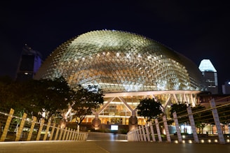 A large, modern architectural structure with a unique dome-like geometric design, illuminated at night. The building is surrounded by trees and pathways, with the city skyline visible in the background.