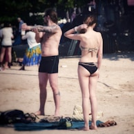 Two individuals in swimwear stand on a sandy beach. One person appears to be applying sunscreen or lotion, while the other stands with their back to the camera. The area is populated with other beachgoers in the background, and there are various bags and towels on the ground.